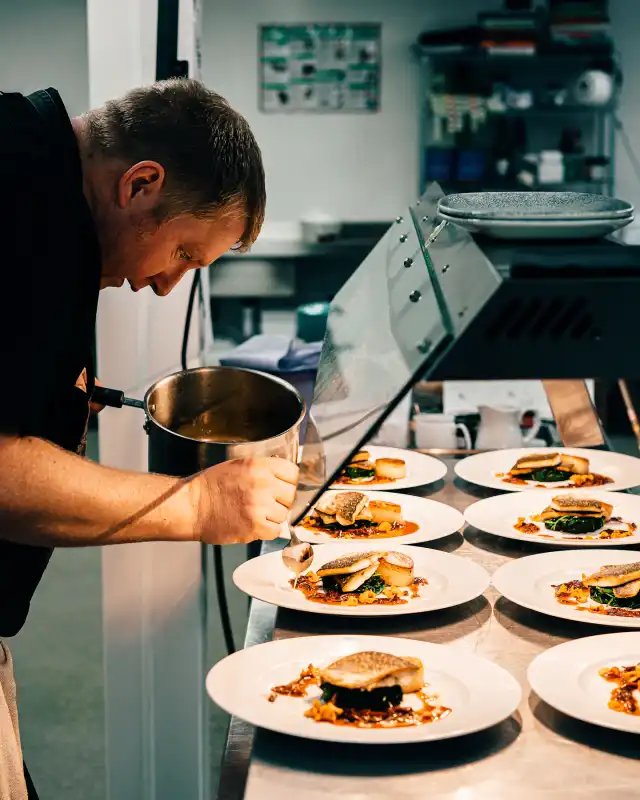 A chef spooning sauce onto plated fish dishes at the pass